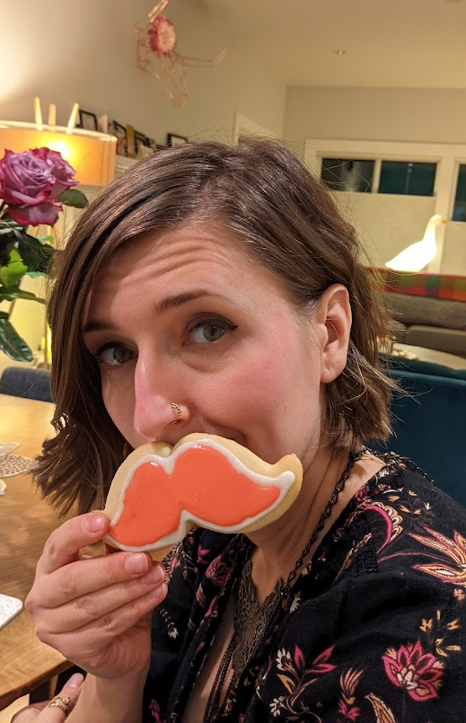 The author (a woman with short hair) is smiling to the camera while holding a mustache shaped cookie up to her face. 